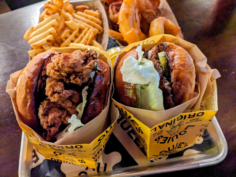 Two burgers wrapped in paper, one with fried chicken and the other with beef, lettuce, and pickles. Served with waffle fries and onion rings on a tray.