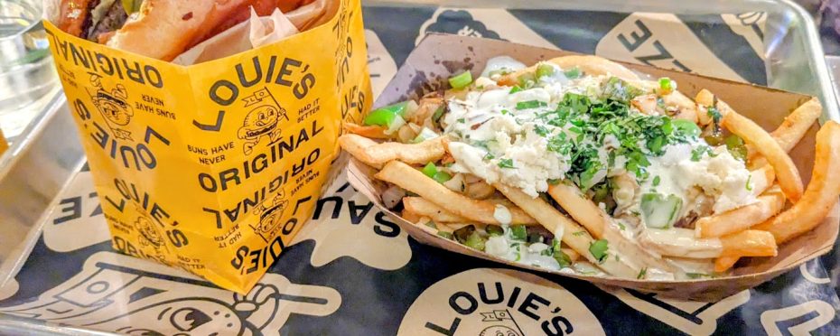 Burger wrapped in yellow paper and loaded fries topped with cheese, sauce, and herbs on a tray with branded paper.