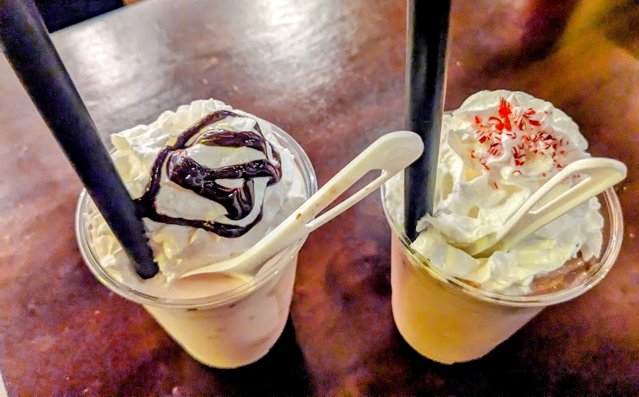 Two milkshakes on a table, one with chocolate syrup and whipped cream, the other with whipped cream and red sprinkles. Each has a spoon and a black straw.
