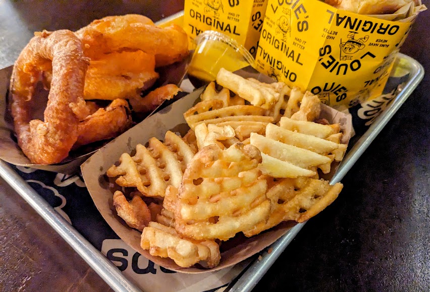 A tray with waffle fries, onion rings, a yellow dipping sauce, and soda cups in yellow branded sleeves.