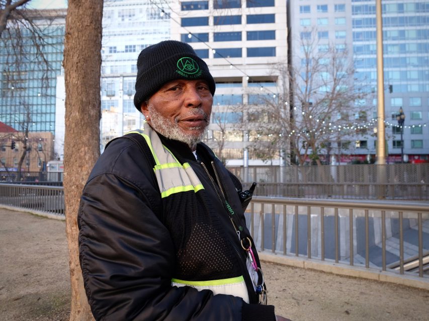 An older man in a black beanie and reflective jacket stands outdoors near a tree, with modern buildings and a railing in the background.