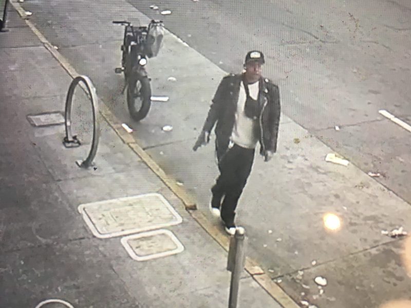 A person in a leather jacket walks confidently on the sidewalk next to a parked motorcycle, as if ready for an action-packed shooting scene.