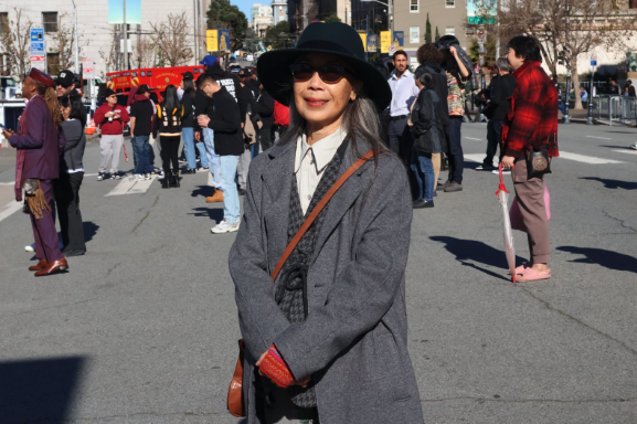 A person in a gray coat and hat stands on a sunny street during an outdoor event. People and emergency vehicles are in the background.