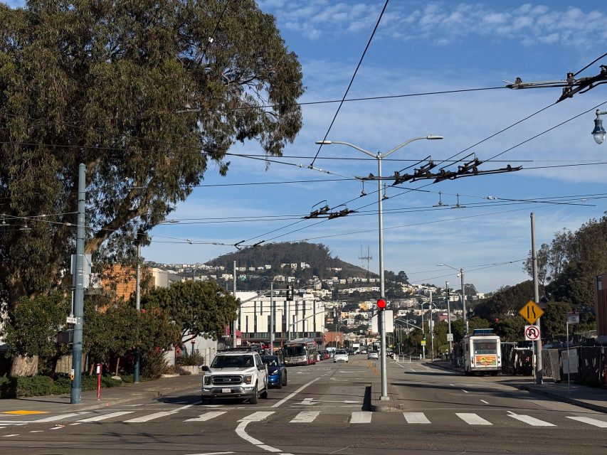 Urban street scene with cars and a bus at an intersection, power lines above, and a hilly residential area in the background under a blue sky.