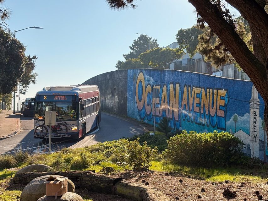 City bus turning on a road near a mural with "Ocean Avenue" painted in large letters, surrounded by trees and greenery.