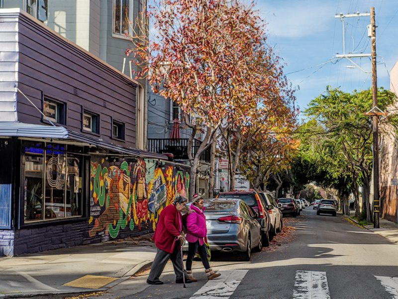 Two people cross a street in an urban area. One uses a cane. Buildings with colorful murals line the sidewalk. A tree with autumn leaves stands nearby. Cars are parked along the street.