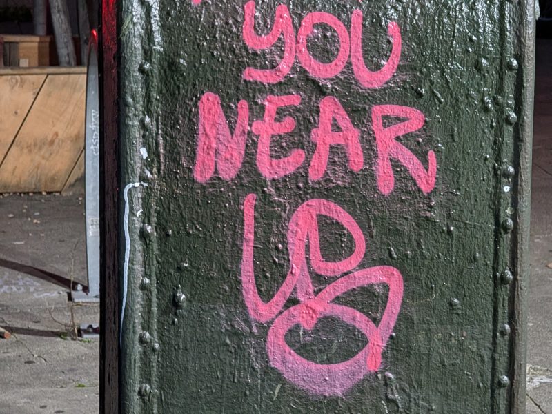 A green mailbox is sprayed with pink graffiti that reads, "HAPPY YOU NEAR UP." It's situated on a sidewalk.
