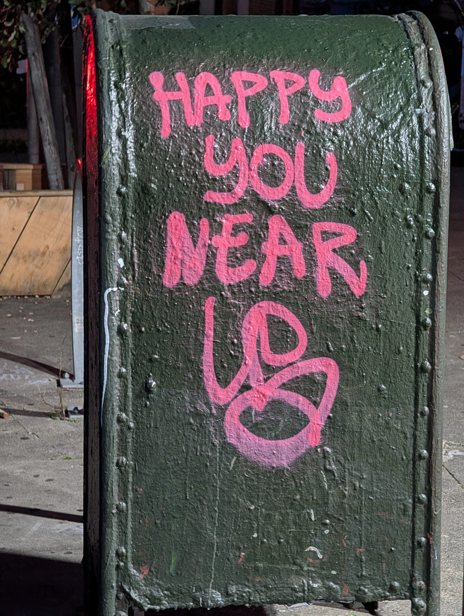 A green mailbox is sprayed with pink graffiti that reads, "HAPPY YOU NEAR UP." It's situated on a sidewalk.