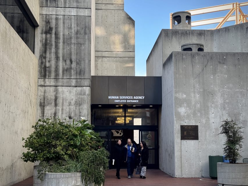 Entrance to the Human Services Agency employee entrance with three people walking out, surrounded by concrete structures and a potted plant.