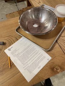 A wooden table with a metal bowl containing dry ingredients, a recipe sheet with handwritten notes, a pencil, and a plastic container with a yellow substance.