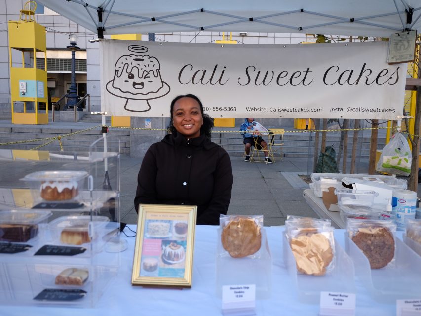 A smiling person stands behind a bakery stall with packaged goods and a "Cali Sweet Cakes" banner featuring contact information.
