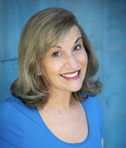 Smiling person with shoulder-length hair wearing a blue shirt against a blue background.