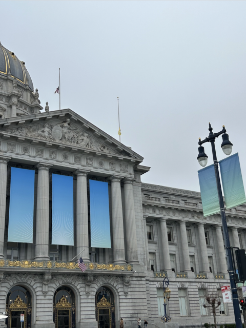 Exterior of a neoclassical building with large columns and a decorative facade. A flag is at half-mast, and banners hang from a lamppost. Overcast sky.
