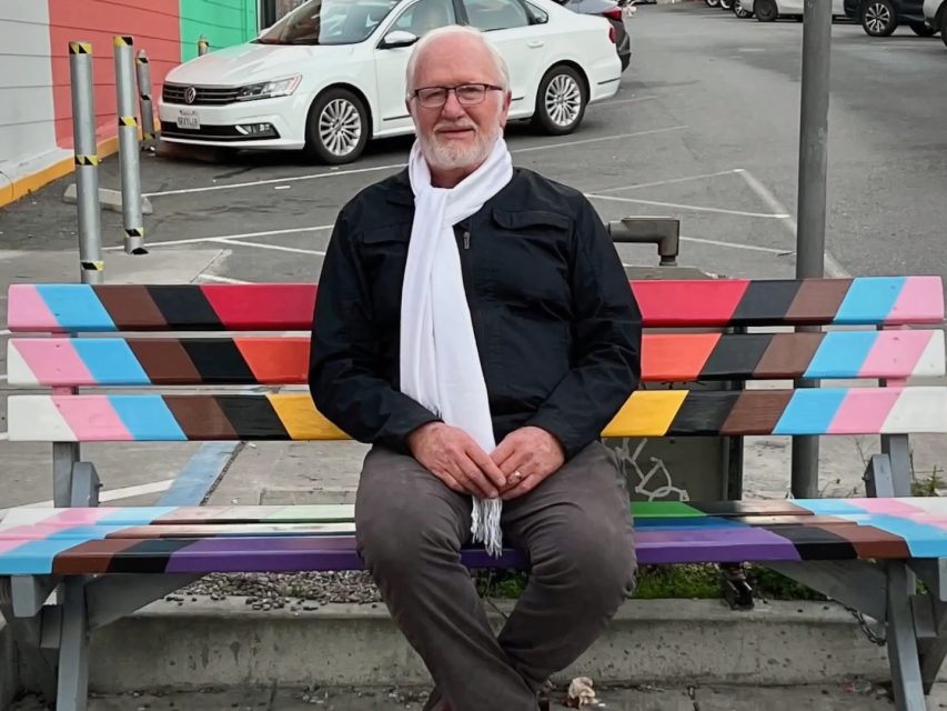 Man sitting on a colorful bench in a parking lot. He's wearing glasses, a white scarf, black jacket, and gray pants. A white car is parked nearby.