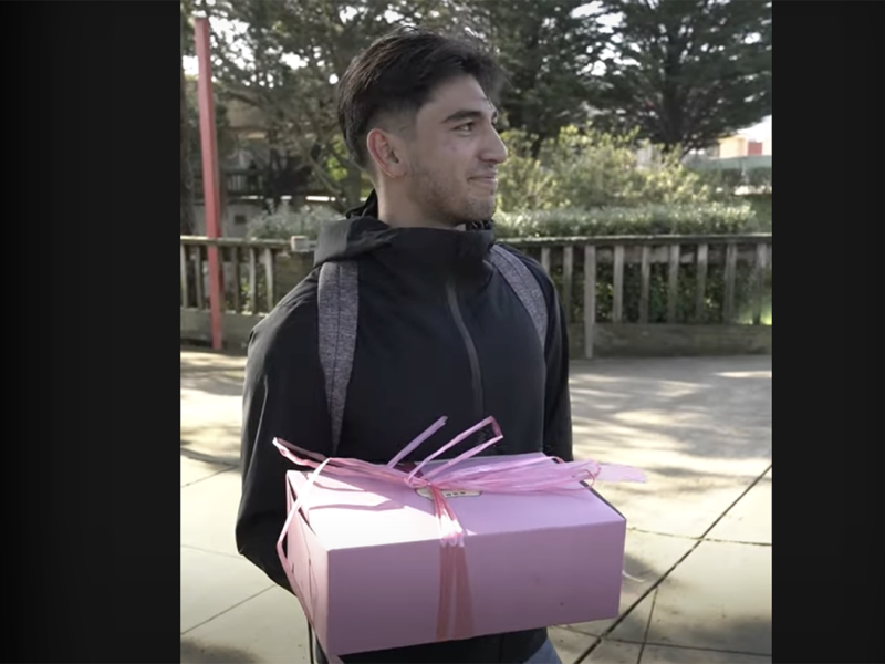 A man in a black jacket holds a pink gift box with ribbons. He is outside, with trees and a fence in the background.