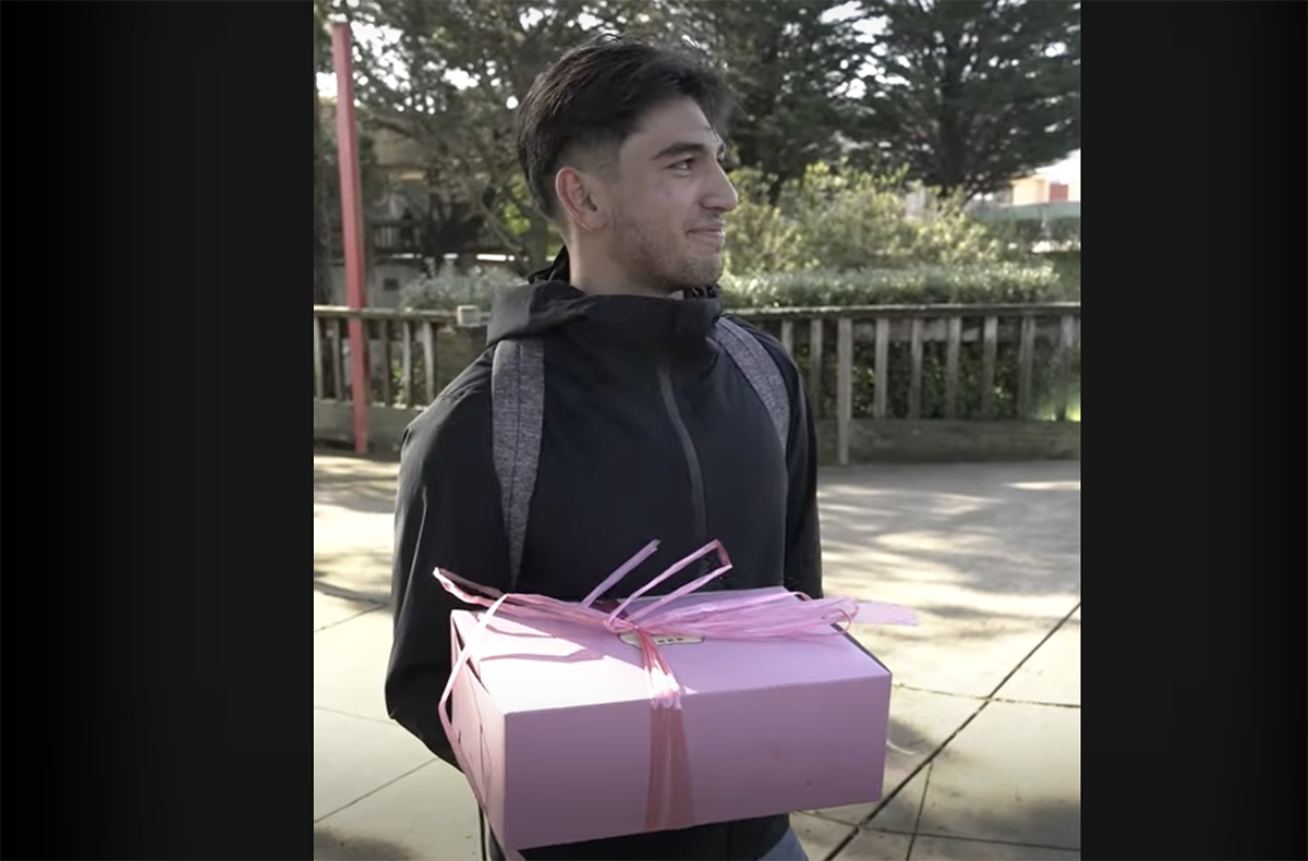 A man in a black jacket holds a pink gift box with ribbons. He is outside, with trees and a fence in the background.