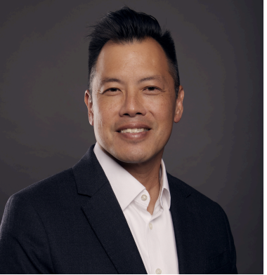 A man in a dark suit and white shirt is smiling against a dark background.