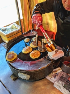Person cooking pancakes on a round griddle using tongs, with a box of cloths nearby.