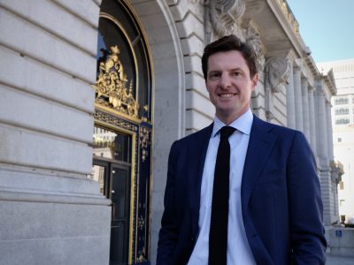 Man in a blue suit and black tie standing in front of an ornate building entrance with columns.
