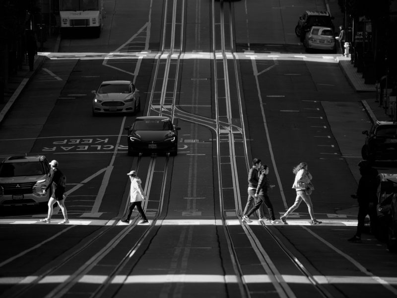 People crossing a street at a crosswalk with a steep incline in the background, flanked by cars and a tram. Black and white photo.