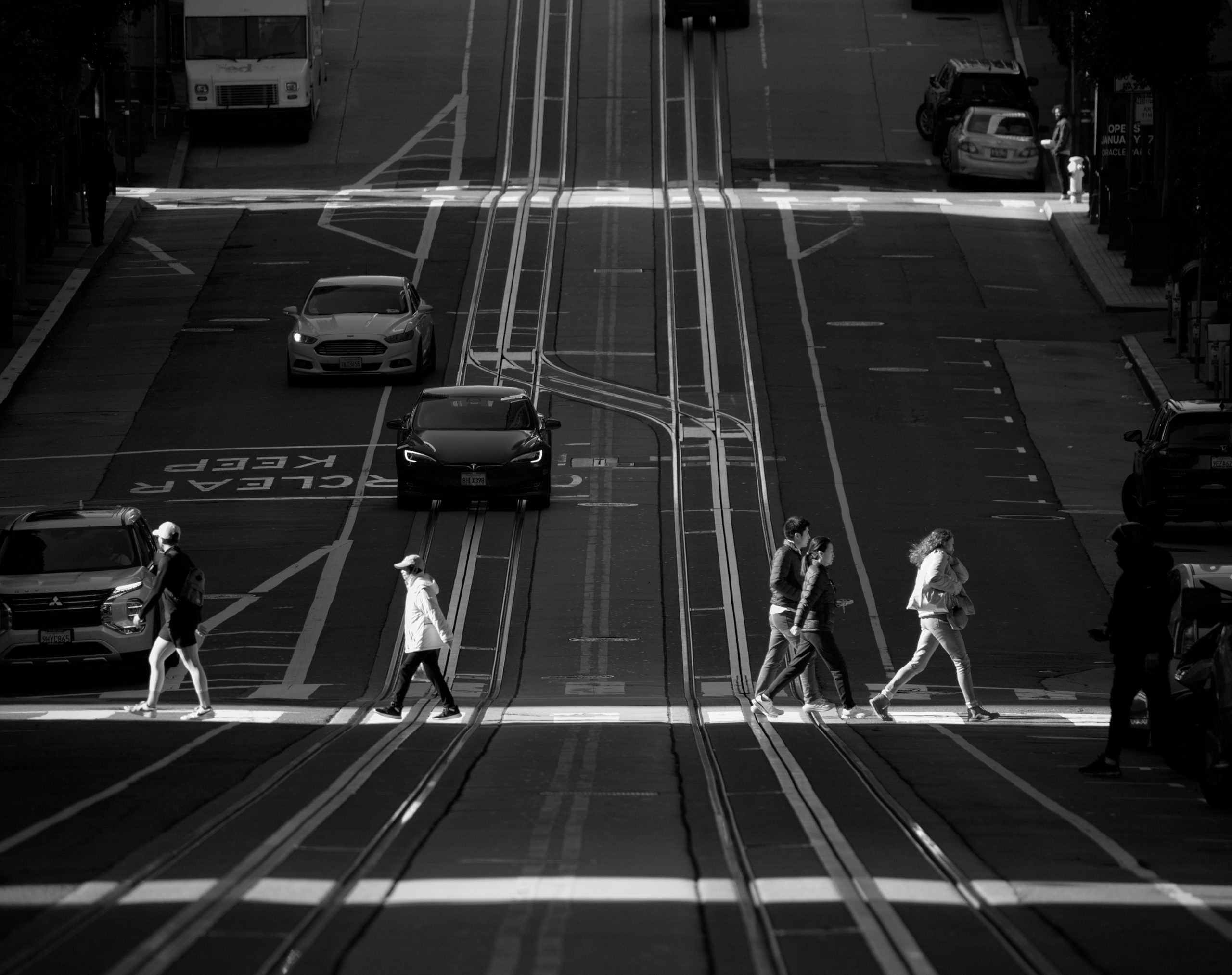 People crossing a street at a crosswalk with a steep incline in the background, flanked by cars and a tram. Black and white photo.