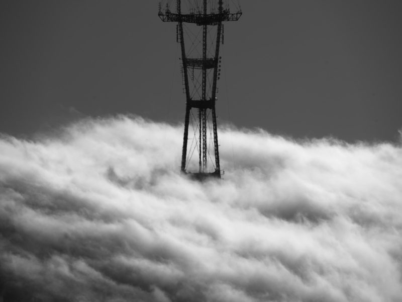 Sutro Tower partially obscured by thick fog, emerging above the cloud layer against a grey sky.