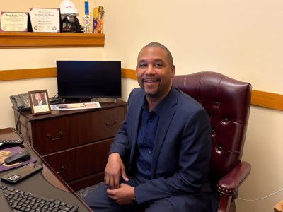 A man in a suit is sitting at a desk in an office. He is smiling, and there is a computer, phone, and framed pictures on the desk. Diplomas and trophies are in the background.
