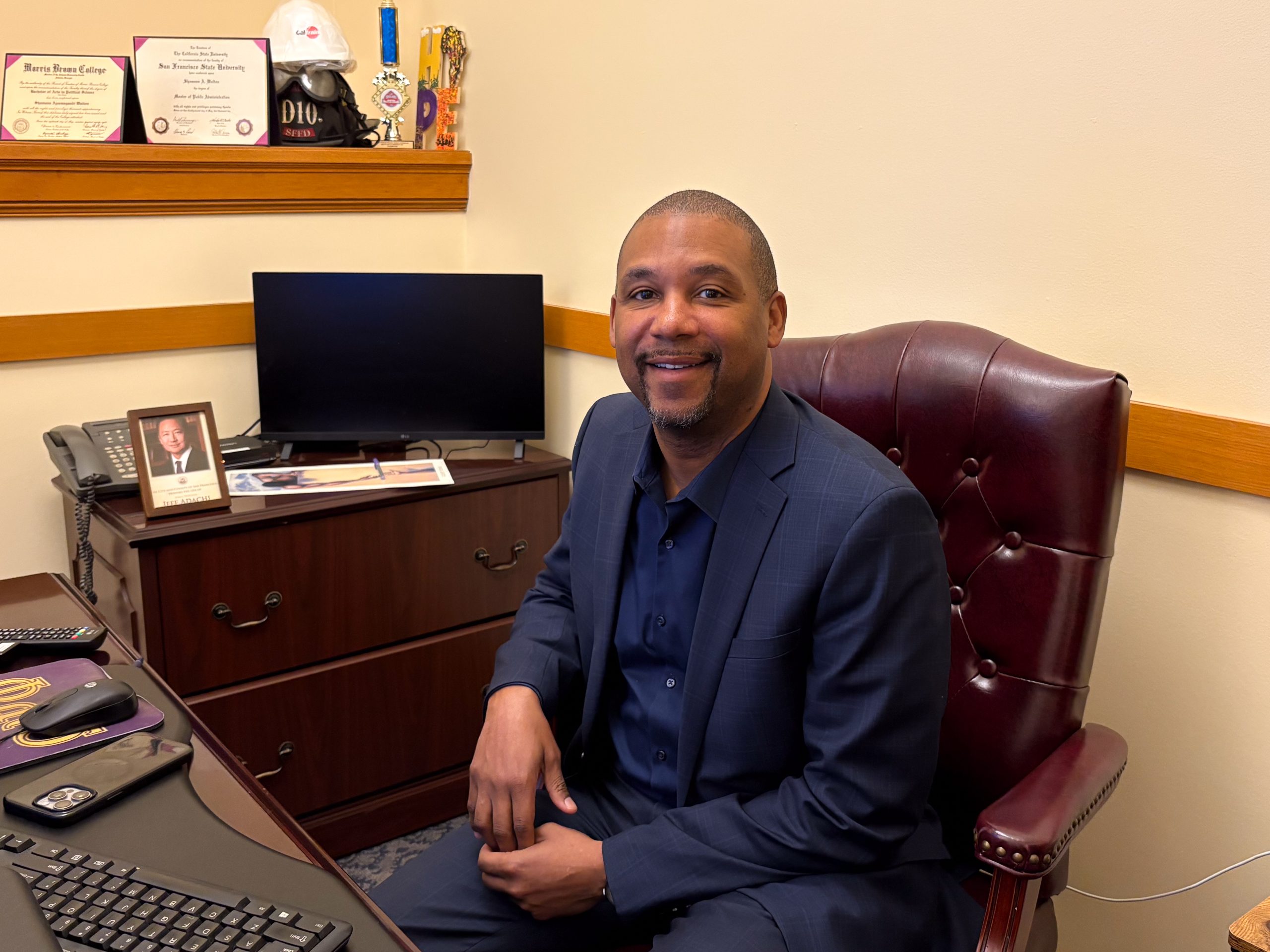 A man in a suit is sitting at a desk in an office. He is smiling, and there is a computer, phone, and framed pictures on the desk. Diplomas and trophies are in the background.