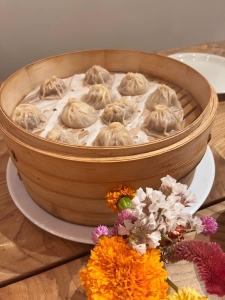 A bamboo steamer with several soup dumplings, resting on a white plate. Flowers are placed in the foreground on a wooden table.