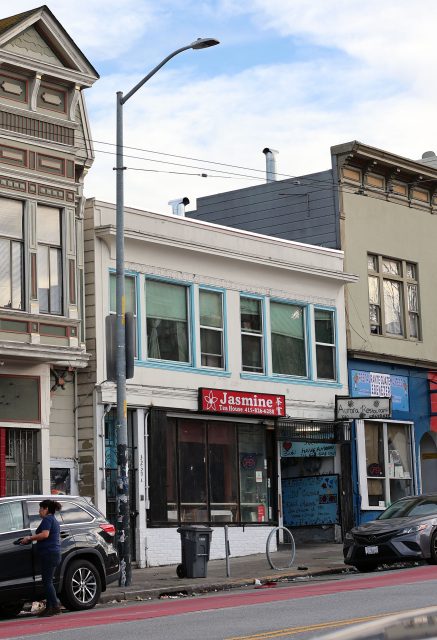 A street view of a two-story building with a "Jasmine" shop on the ground floor. The building has large windows and a garbage bin on the sidewalk. A person stands near a car in the foreground.