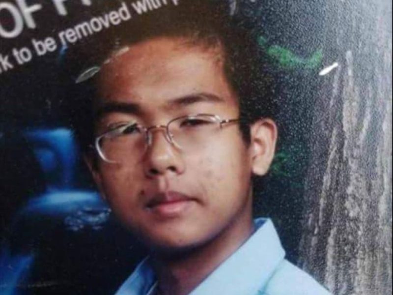 A young person with short dark hair and glasses looks at the camera, wearing a light blue shirt. The background contains indistinct shooting text and some greenery.