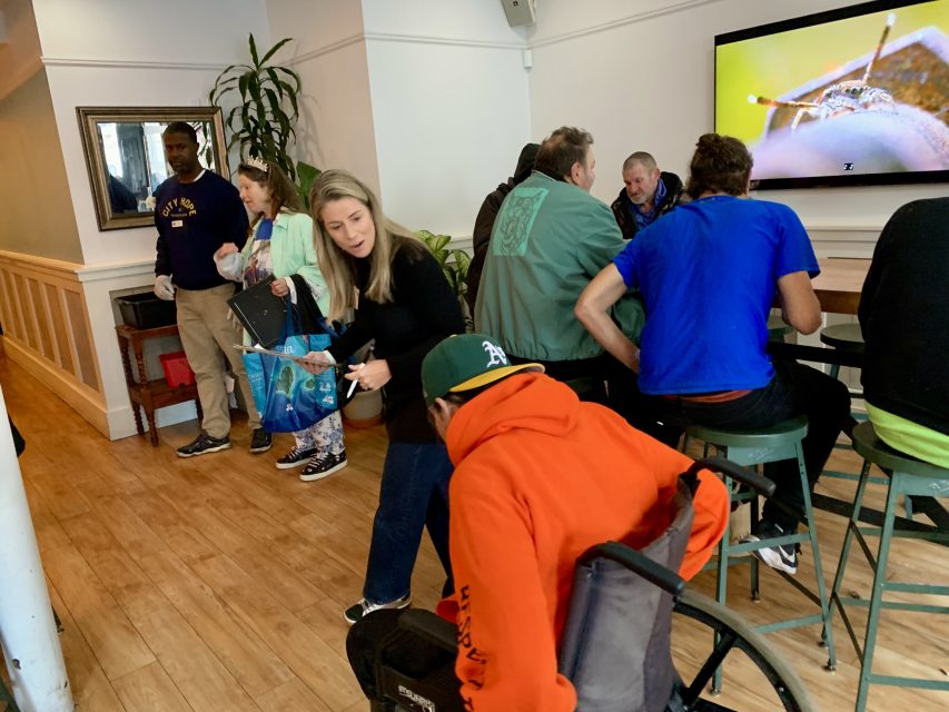 People seated and standing in a cafe or waiting area, engaging in conversation. A TV screen displays a close-up image of a snail.