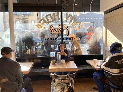 People sitting inside a cafe near the window, with pedestrians visible outside. The cafe's name is displayed on the glass.
