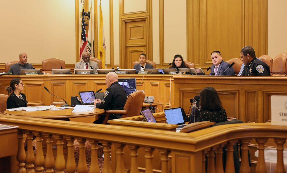 A government meeting in a courtroom setting with officials seated at a panel. A police officer and other individuals are present, with laptops and documents on the table. Flags are in the background.