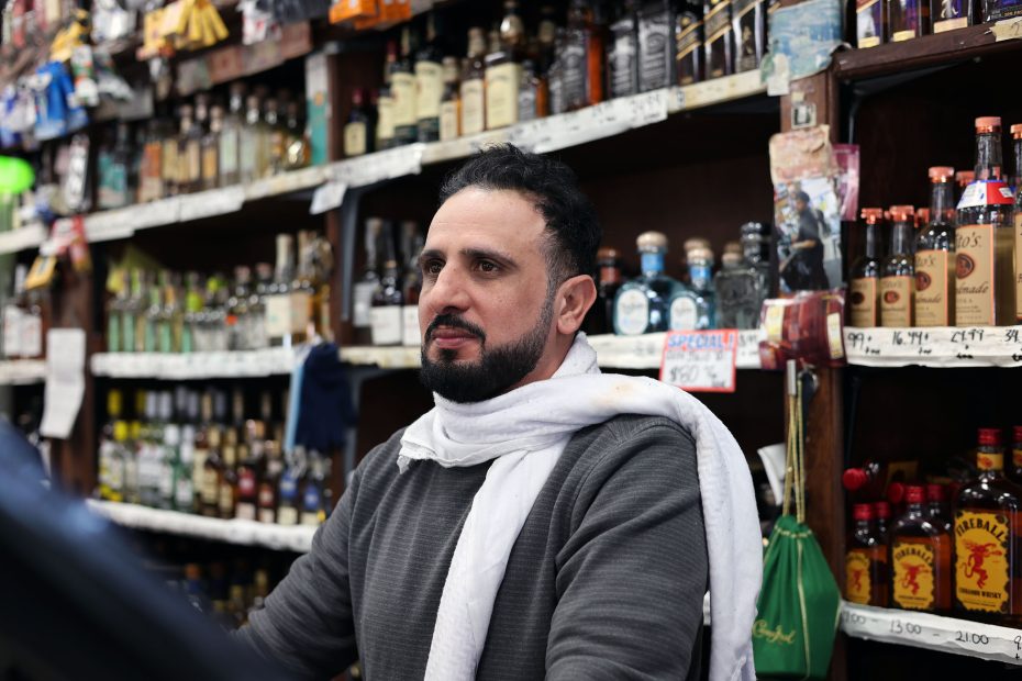 A man stands in a store with shelves of various liquor bottles behind him, quietly observing as customers make their choices, unaware of the shooting excitement unfolding in the neighborhood.