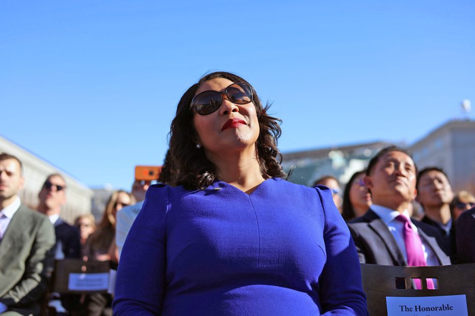 A woman in a blue dress and sunglasses sits among a group of people outdoors under a clear sky.