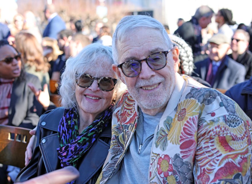 An elderly couple sitting together outdoors, smiling at the camera. The man is wearing glasses and a colorful jacket, while the woman has sunglasses and a leather jacket. Other people are around them.