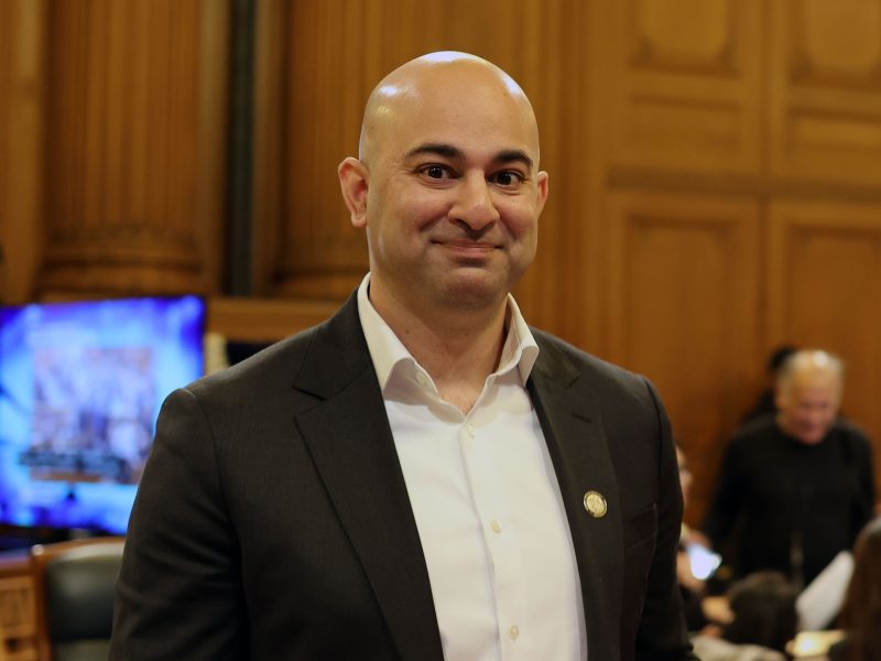 A person in a suit stands in a wood-paneled room with chairs and a screen in the background.