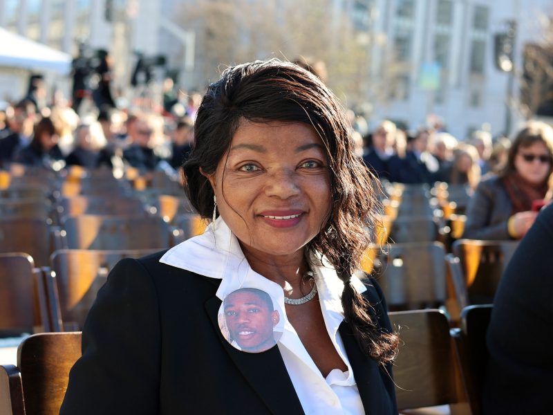 A woman sits outdoors on a wooden chair, wearing a black blazer, white shirt, and a pin with a young man's photo. Crowds and trees are visible in the background.