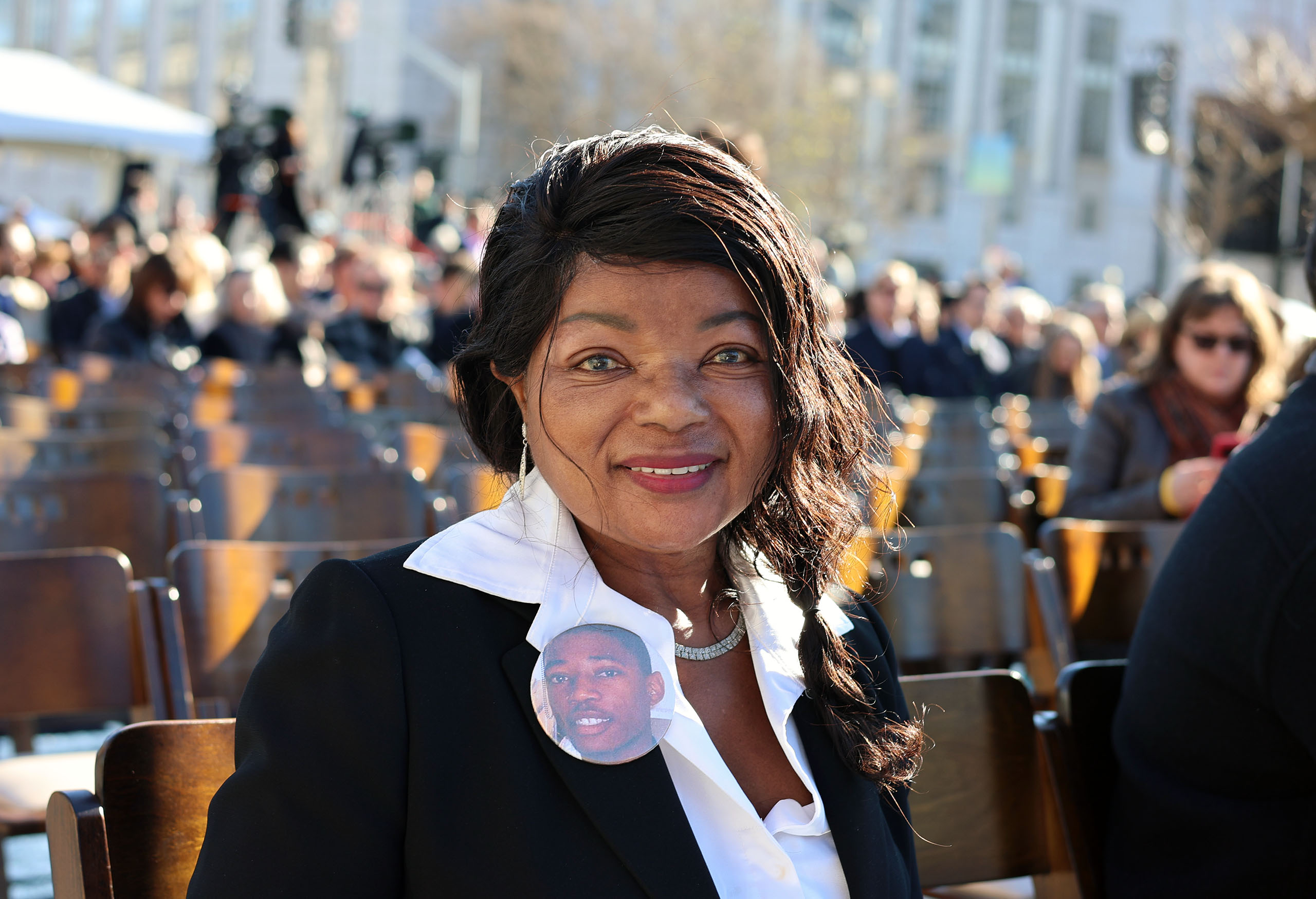 A woman sits outdoors on a wooden chair, wearing a black blazer, white shirt, and a pin with a young man's photo. Crowds and trees are visible in the background.