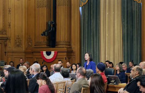 A person stands speaking in a wood-paneled room filled with seated people. The room has curtains and a small red, white, and blue decoration on the wall.