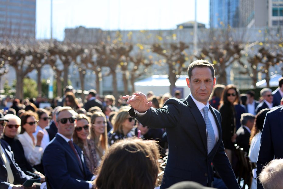 A man in a suit extends his hand while walking through a seated crowd outdoors, with trees and buildings in the background.