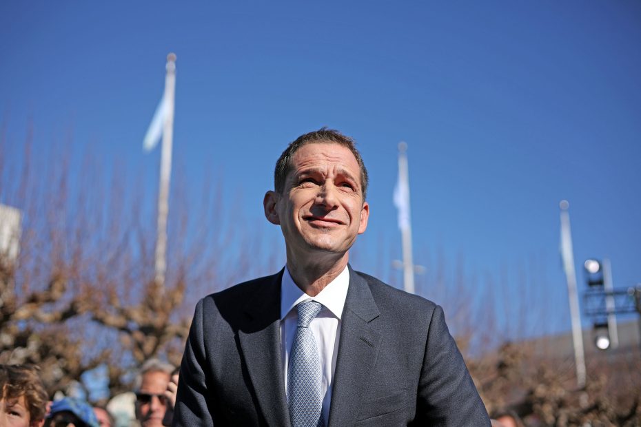 A man in a suit and tie stands outdoors under a blue sky, looking upward. People and bare trees are in the background.