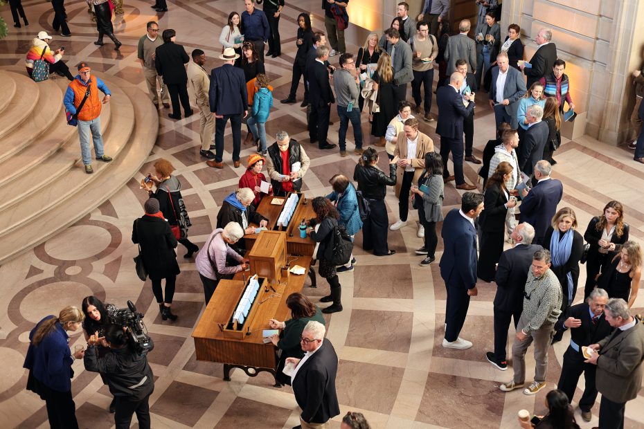 A large group of people gathers in a spacious hall with patterned flooring. Several individuals are signing documents at tables in the center.
