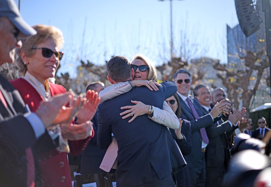 A man and woman hug surrounded by applauding people outdoors.