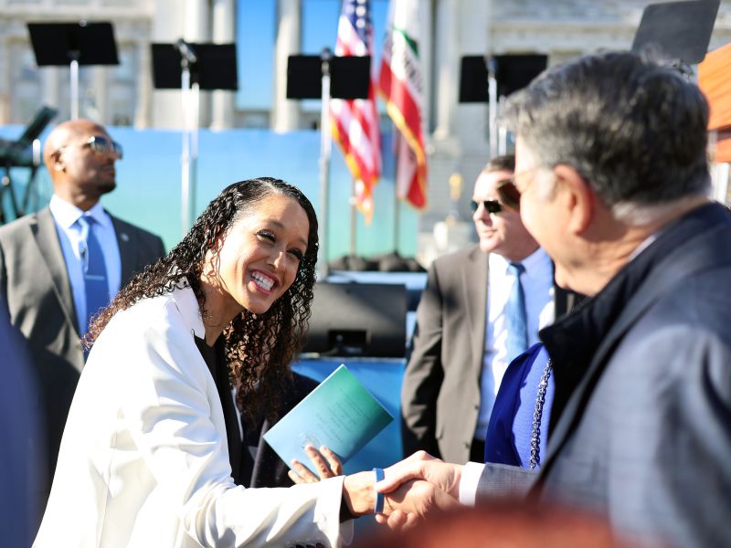 Woman in a white blazer shakes hands with a man at an outdoor event. American flag and stage in the background.