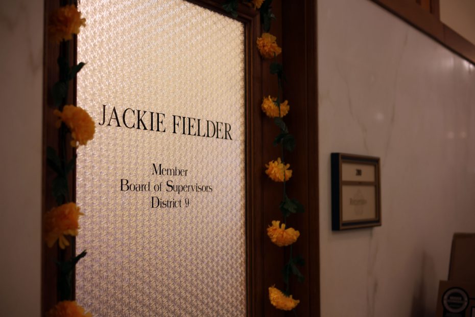An office door with the name "Jackie Fielder, Member, Board of Supervisors, District 9" and yellow flower decorations on the frame.