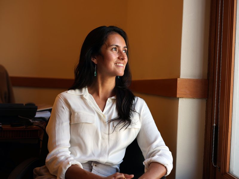 Jackie Fielder in a white shirt sits on a chair by a window, smiling and looking outside.