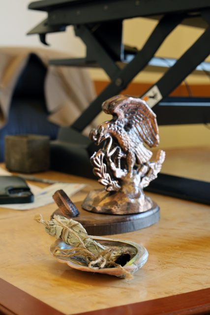 Desk with a bronze eagle statue and a small bowl containing dried sage leaves. An adjustable stand and a folded coat are in the background.