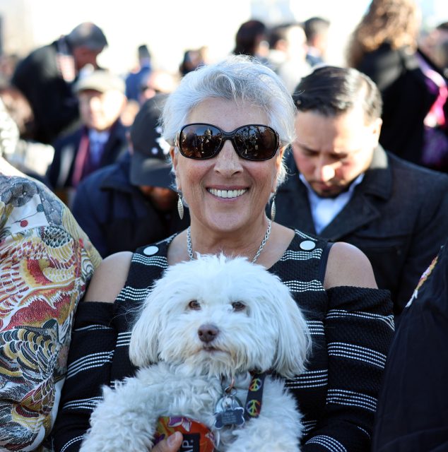 A woman with sunglasses smiles while holding a white dog on her lap in a crowded outdoor setting.